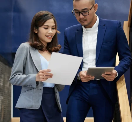 Dua profesional muda berdiskusi sambil memegang dokumen dan tablet, merepresentasikan kerja tim bersama ICF-Certified Coaches.